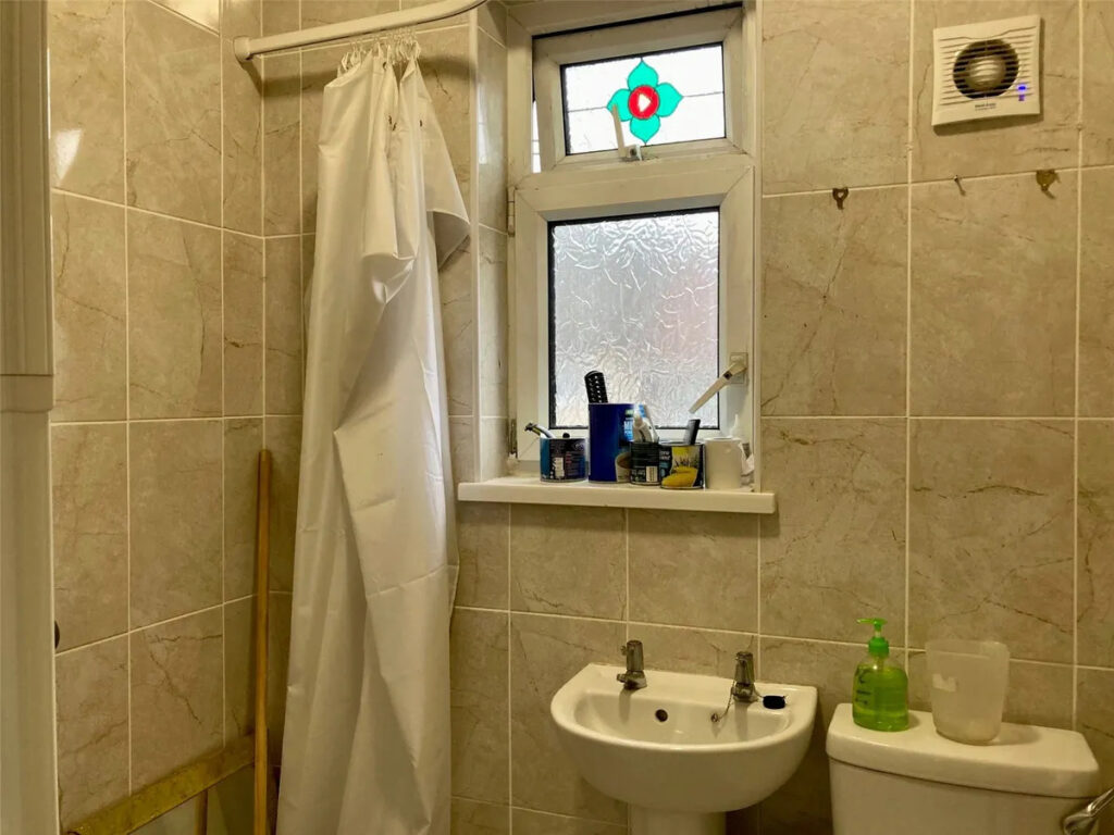 Property for sale A small tiled bathroom in a Stanley 3-bed terrace, featuring a shower curtain, frosted window with colorful design, twin-tap sink, toiletries on the sill, soap dispenser, and wall-mounted extractor fan—perfect for a County Durham house auction. Presented by Palace Auctions