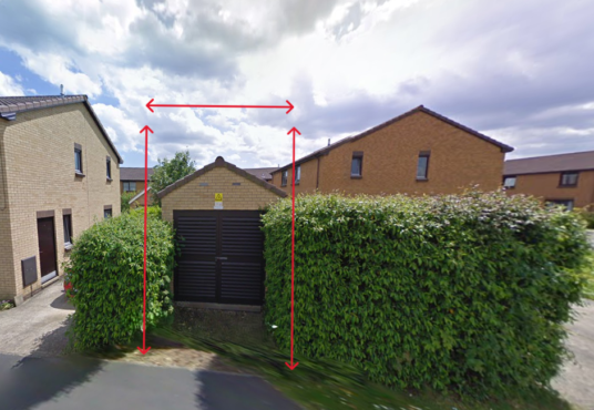 Property for sale A brown garage with black doors sits between green hedges, marked by red arrows showing its height and width—an ideal alternative real estate asset. Houses and a street appear in the background under a cloudy sky. Presented by Palace Auctions