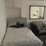 Property for sale A small bedroom in Gwydyr Mansions Brighton features an unmade bed against a white wall, pillows and a blanket, a beige sofa to the right, a large mirror reflecting another room, and two closed cabinets above the bed. Presented by Palace Auctions