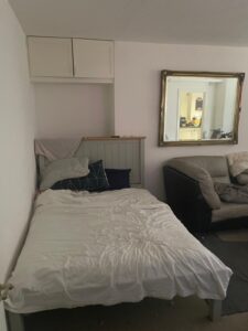 Property for sale A small bedroom in Gwydyr Mansions Brighton features an unmade bed against a white wall, pillows and a blanket, a beige sofa to the right, a large mirror reflecting another room, and two closed cabinets above the bed. Presented by Palace Auctions