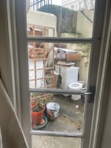 Property for sale View through a window reveals a cluttered outdoor area at Gwydyr Mansions Brighton, with a washing machine, boxes, wooden items, potted plant, and debris on concrete. Stairs and railings are seen—potential investment property Brighton opportunity. Presented by Palace Auctions