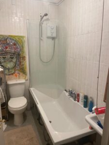 Property for sale A small bathroom in Gwydyr Mansions Brighton features a bathtub, wall-mounted shower, toilet, and toiletries on the tub edge. Above the toilet hangs a colorful painting of a person in a headdress. A towel and mat are also visible. Presented by Palace Auctions