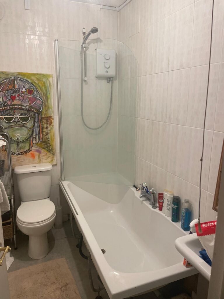 Property for sale A small bathroom in Gwydyr Mansions Brighton features a bathtub, wall-mounted shower, toilet, and toiletries on the tub edge. Above the toilet hangs a colorful painting of a person in a headdress. A towel and mat are also visible. Presented by Palace Auctions