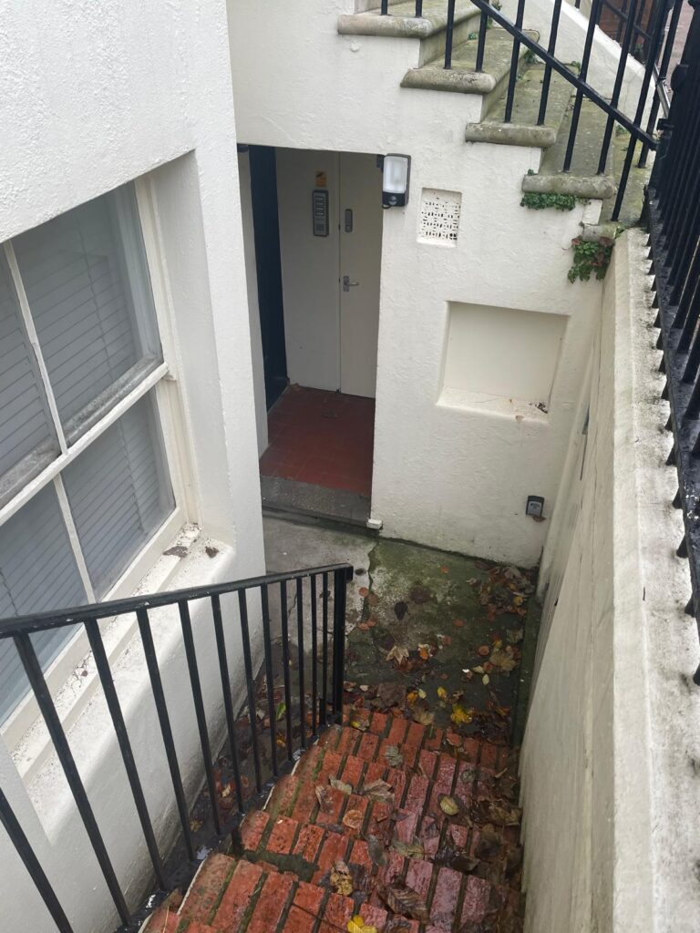 Property for sale A narrow brick staircase with black railings leads down to a basement entrance of a white building—an ideal investment property Brighton seekers will appreciate. The ground is damp with scattered leaves, and a door is visible at the bottom. Presented by Palace Auctions