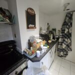 Property for sale A small kitchen in a Hove apartment for sale, with white cabinets and cluttered countertops holding groceries, bottles, and kitchen items. A plaid cloth hangs on the wall alongside a Coca-Cola chalkboard, while a white tile floor completes this investment property. Presented by Palace Auctions
