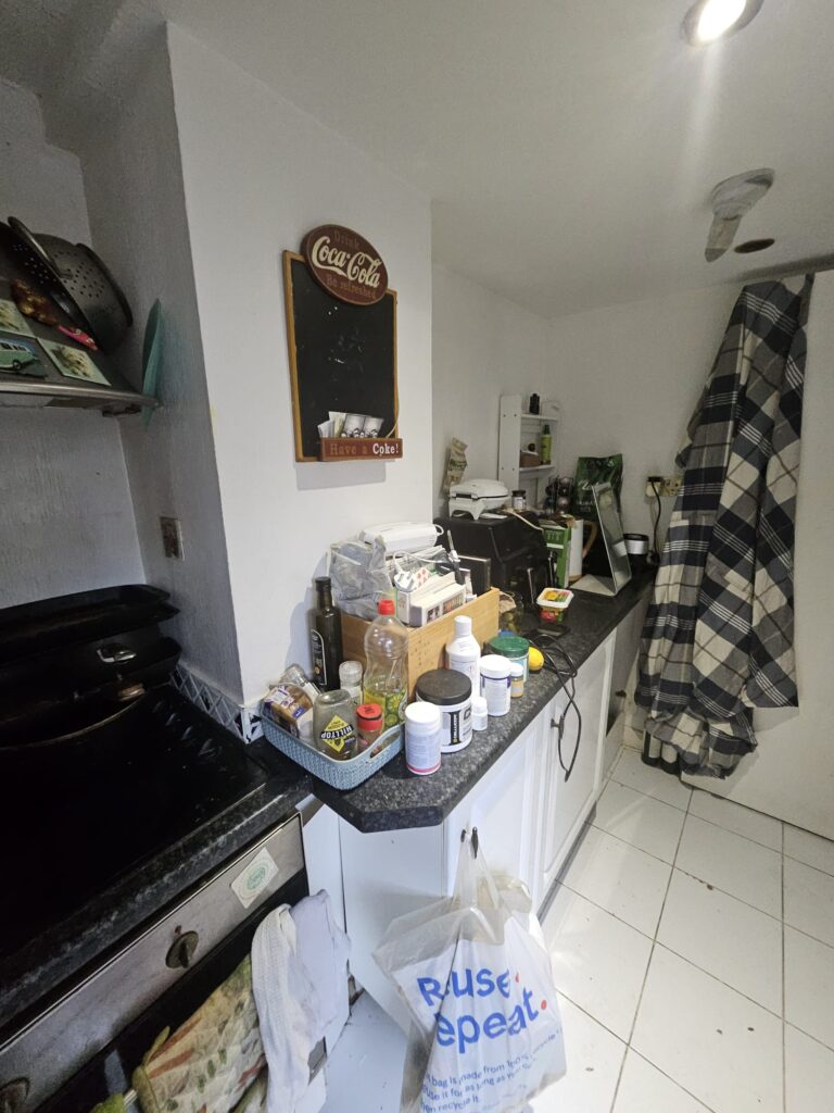 Property for sale A small kitchen in a Hove apartment for sale, with white cabinets and cluttered countertops holding groceries, bottles, and kitchen items. A plaid cloth hangs on the wall alongside a Coca-Cola chalkboard, while a white tile floor completes this investment property. Presented by Palace Auctions