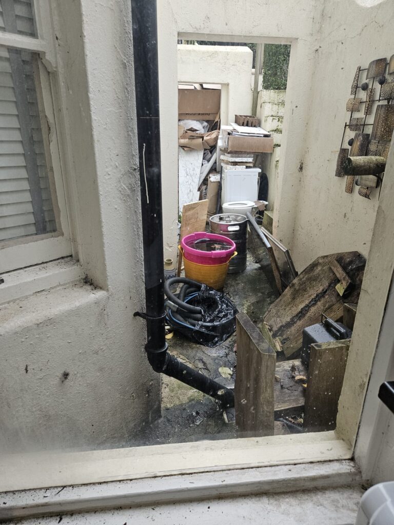 Property for sale A cluttered outdoor area viewed through a window in a Hove apartment for sale, filled with buckets, hoses, wooden planks, and a white appliance, all surrounded by dirty, worn walls and assorted debris. Presented by Palace Auctions