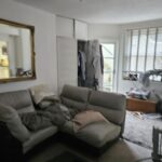 Property for sale A living room in one of the stylish flats for sale Brighton, featuring a beige sectional sofa, cozy pillows and blankets, a large mirror, an open closet with clothes, blinds on the window, and assorted items on the floor and surfaces. Presented by Palace Auctions