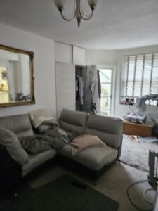 Property for sale A living room in one of the stylish flats for sale Brighton, featuring a beige sectional sofa, cozy pillows and blankets, a large mirror, an open closet with clothes, blinds on the window, and assorted items on the floor and surfaces. Presented by Palace Auctions