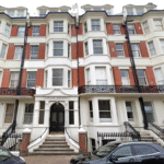Property for sale A four-story Victorian-style apartment building with red brick and white trim, arched entrance labeled "Olinda Mansions," iron railings, bay windows, and black cars parked in front—ideal for those seeking unique flats for sale Brighton. Presented by Palace Auctions