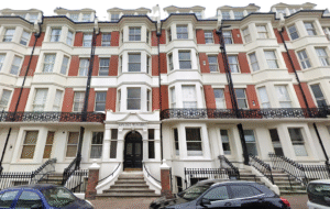 Property for sale A four-story Victorian-style apartment building with red brick and white trim, arched entrance labeled "Olinda Mansions," iron railings, bay windows, and black cars parked in front—ideal for those seeking unique flats for sale Brighton. Presented by Palace Auctions