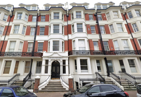 Property for sale A four-story Victorian-style apartment building with red brick and white trim, arched entrance labeled "Olinda Mansions," iron railings, bay windows, and black cars parked in front—ideal for those seeking unique flats for sale Brighton. Presented by Palace Auctions