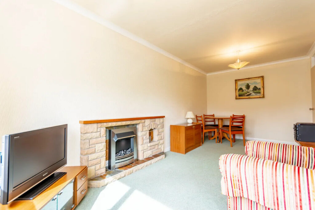 Property for sale A bright living room in this one bedroom flat Nottingham features a TV on a stand, stone fireplace, striped armchair, and a small dining area with wooden chairs and table. A painting hangs on the wall above the dining area. Presented by Palace Auctions