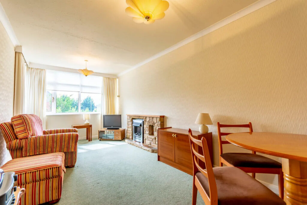 Property for sale Bright living room in a one-bedroom flat Nottingham featuring a striped armchair, small wooden dining table, TV on a stand, fireplace, wooden sideboard, and a large window filling the space with natural light. Presented by Palace Auctions