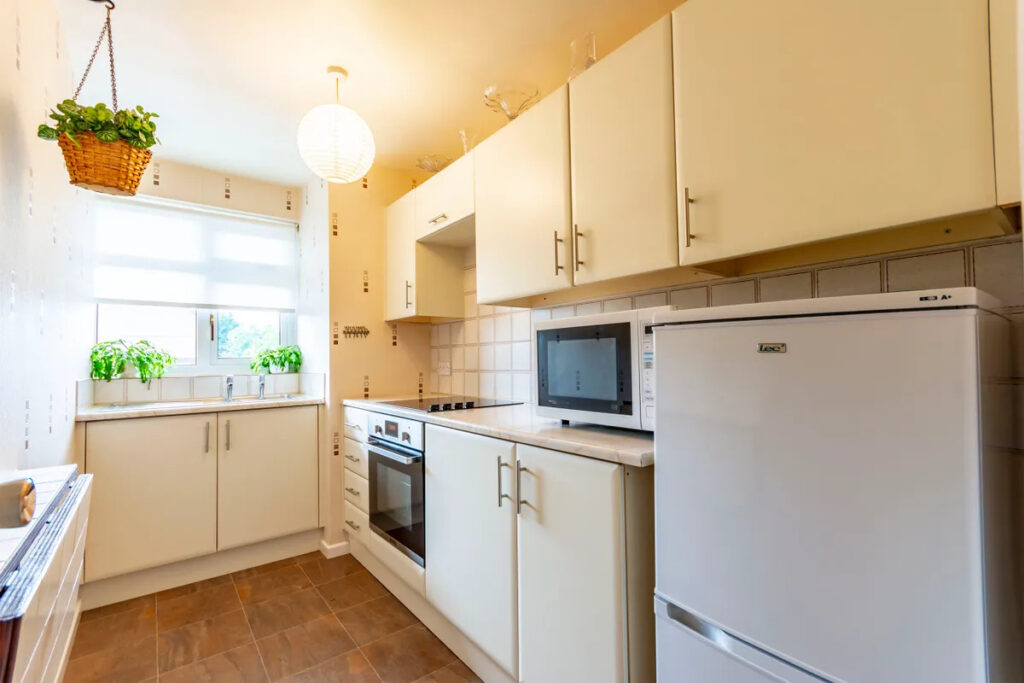 Property for sale Bright kitchen with white cabinets, built-in oven, microwave, and fridge in a one-bedroom flat Nottingham; potted plants on the windowsill and hanging from the ceiling fill the space with charm and natural light from a large window. Presented by Palace Auctions
