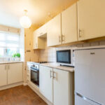Property for sale Bright kitchen with white cabinets, built-in oven, microwave, and fridge in a one-bedroom flat Nottingham; potted plants on the windowsill and hanging from the ceiling fill the space with charm and natural light from a large window. Presented by Palace Auctions