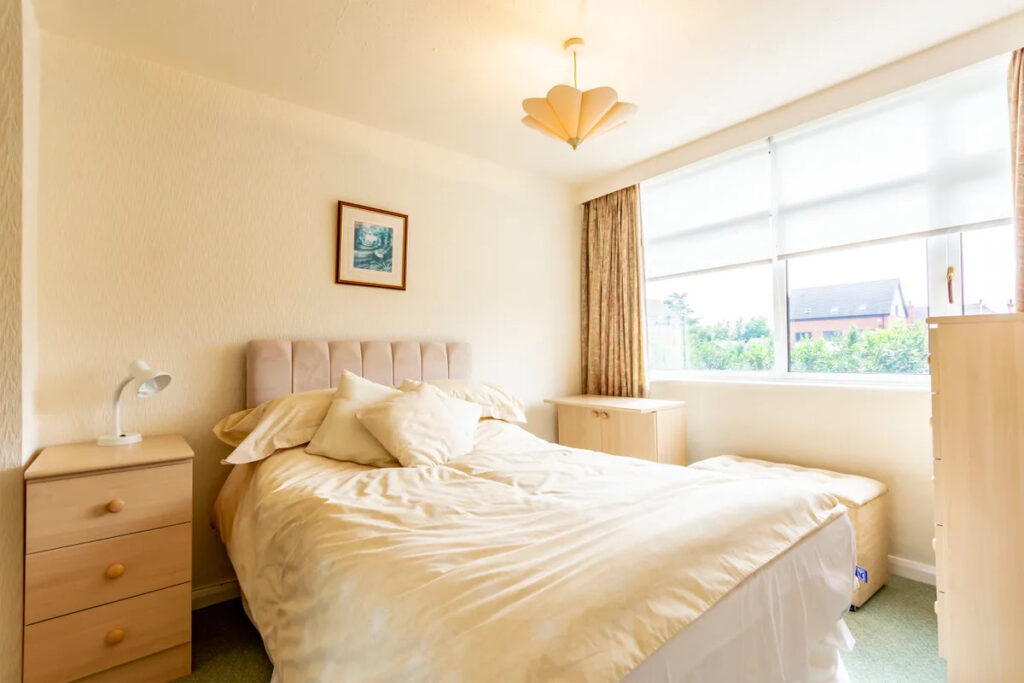 Property for sale A bright bedroom with a double bed, light bedding, wooden nightstands, a dresser, and a large window with beige curtains—ideal for a buy-to-let West Bridgford investor seeking comfortable living spaces filled with natural light. Presented by Palace Auctions