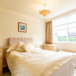 Property for sale A bright bedroom with a double bed, light bedding, wooden nightstands, a dresser, and a large window with beige curtains—ideal for a buy-to-let West Bridgford investor seeking comfortable living spaces filled with natural light. Presented by Palace Auctions