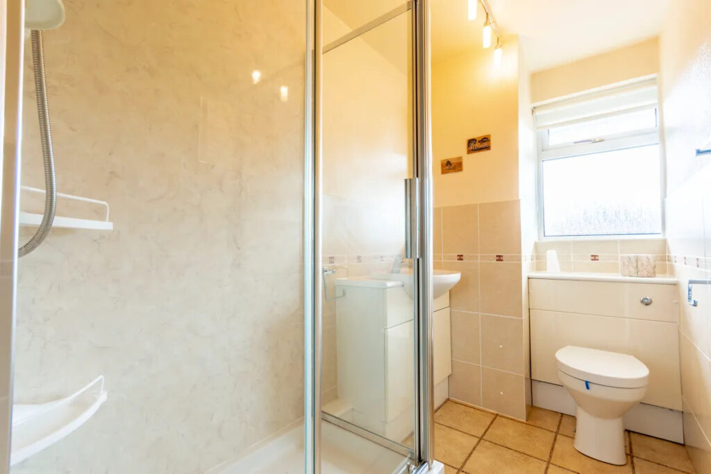 Property for sale Bright bathroom in a one-bedroom flat Nottingham, featuring a glass shower enclosure, beige tiled floors, a white toilet, a small sink with cabinet, and a window letting in natural light. The walls are light-colored and minimally decorated. Presented by Palace Auctions