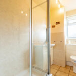 Property for sale Bright bathroom in a one-bedroom flat Nottingham, featuring a glass shower enclosure, beige tiled floors, a white toilet, a small sink with cabinet, and a window letting in natural light. The walls are light-colored and minimally decorated. Presented by Palace Auctions