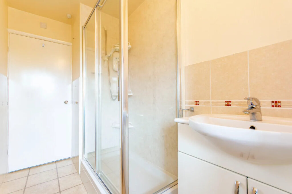 Property for sale A bright bathroom in a one-bedroom flat Nottingham featuring a glass shower enclosure, white sink with cabinet, beige tiled walls, and a closed white door in the background. Presented by Palace Auctions
