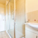 Property for sale A bright bathroom in a one-bedroom flat Nottingham featuring a glass shower enclosure, white sink with cabinet, beige tiled walls, and a closed white door in the background. Presented by Palace Auctions