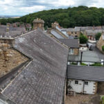 Property for sale A view over the rooftops of a small village near Newcastle upon Tyne, with stone houses and narrow streets—perfect for a traditional public house conversion in the Newcastle area—set against a forested hill and partly cloudy sky. Presented by Palace Auctions