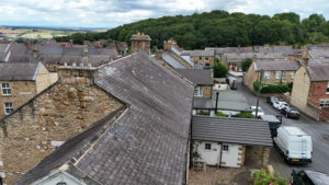 Property for sale A view over the rooftops of a small village near Newcastle upon Tyne, with stone houses and narrow streets—perfect for a traditional public house conversion in the Newcastle area—set against a forested hill and partly cloudy sky. Presented by Palace Auctions