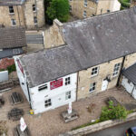 Property for sale Aerial view of a freehold property with beer garden NE16 6NF—a stone building with a gray tiled roof and a red sign reading “The Wheatsheaf Inn,” featuring gravel outdoor seating, parked cars, and neighboring stone buildings. Presented by Palace Auctions
