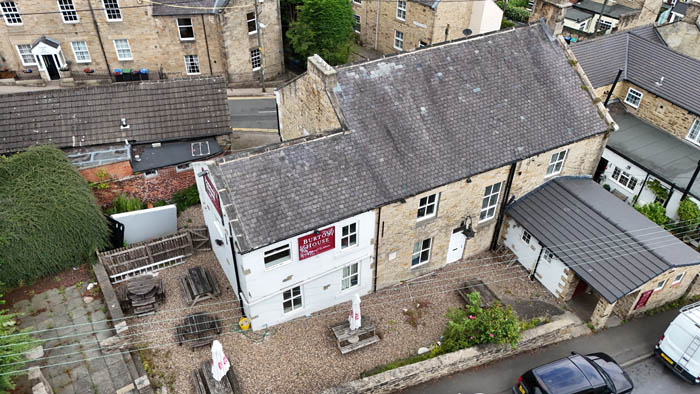 Property for sale Aerial view of a freehold property with beer garden NE16 6NF—a stone building with a gray tiled roof and a red sign reading “The Wheatsheaf Inn,” featuring gravel outdoor seating, parked cars, and neighboring stone buildings. Presented by Palace Auctions