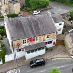 Property for sale Aerial view of a detached public house for sale in Burnopfield, Newcastle upon Tyne, with a red "For Sale" sign at a street corner. Cars are parked nearby, and surrounding buildings and greenery are visible. Presented by Palace Auctions