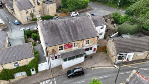 Property for sale Aerial view of a detached public house for sale in Burnopfield, Newcastle upon Tyne, with a red "For Sale" sign at a street corner. Cars are parked nearby, and surrounding buildings and greenery are visible. Presented by Palace Auctions