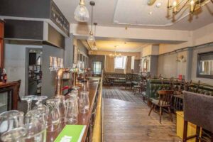Property for sale A traditional public house conversion in the Newcastle area, featuring wooden floors, green upholstered seating, a long bar lined with glassware, and pendant lighting. Natural light fills the room from windows at the back. Presented by Palace Auctions