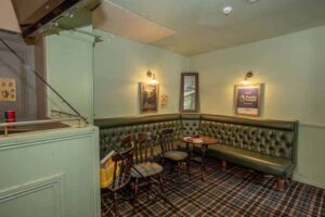 Property for sale A cozy pub corner with green tufted banquette seating, wooden chairs, small round tables, and a plaid carpet. This traditional public house conversion in the Newcastle area features dim wall lights and framed posters, with a highchair tucked beside the seating. Presented by Palace Auctions