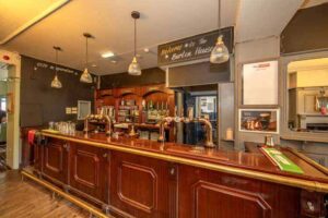 Property for sale A traditional pub bar with wooden counters and beer taps, glassware neatly arranged, pendant lights overhead, and a sign reading “Welcome to The Burton House”—a classic public house conversion in the Newcastle area. Presented by Palace Auctions