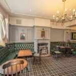 Property for sale A cozy, traditional public house conversion in the Newcastle area, featuring green leather seating, wooden tables and chairs, a fireplace with a small mantel, checkered carpet, and framed pictures on light gray walls under a chandelier. Presented by Palace Auctions