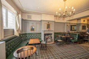 Property for sale A cozy, traditional public house conversion in the Newcastle area, featuring green leather seating, wooden tables and chairs, a fireplace with a small mantel, checkered carpet, and framed pictures on light gray walls under a chandelier. Presented by Palace Auctions