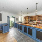 Property for sale A bright, empty pub interior with blue and wood paneling, a wooden bar counter, hanging lights, bar stools, and an open door leading outside—ideal for a traditional public house conversion in the Newcastle area. Presented by Palace Auctions