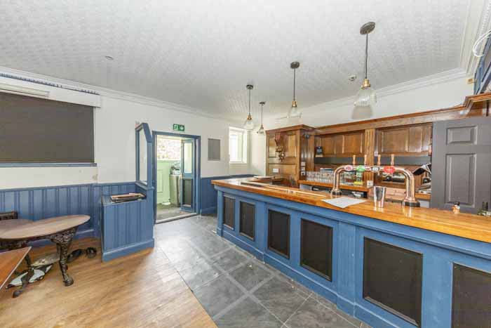 Property for sale A bright, empty pub interior with blue and wood paneling, a wooden bar counter, hanging lights, bar stools, and an open door leading outside—ideal for a traditional public house conversion in the Newcastle area. Presented by Palace Auctions