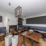 Property for sale A cluttered room with wooden tables and chairs, two white bins, blue paneling, covered windows, and an unlit fireplace hints at a traditional public house conversion in the Newcastle area. The space appears unused or in transition. Presented by Palace Auctions