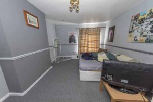 Property for sale A small bedroom with gray walls, patterned bedding, a TV on a wooden table, drying rack, wall art, and a window with brown patterned curtains letting in daylight—ideal for a traditional public house conversion in the Newcastle area. Presented by Palace Auctions