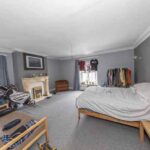 Property for sale A spacious, cluttered bedroom with gray walls, a bed, sofa, fireplace, bike, and clothing rack—ideal for those viewing an auction property for sale in Newcastle upon Tyne. Natural light fills the room through two curtained windows. Presented by Palace Auctions