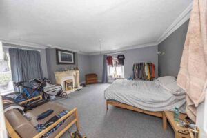 Property for sale A spacious, cluttered bedroom with gray walls, a bed, sofa, fireplace, bike, and clothing rack—ideal for those viewing an auction property for sale in Newcastle upon Tyne. Natural light fills the room through two curtained windows. Presented by Palace Auctions