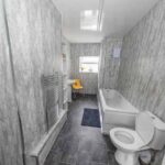 Property for sale A modern bathroom in a traditional public house conversion, featuring gray marble-style walls and floor, a bathtub with shower, white toilet, towel radiator, small window, yellow chair, and blue rug—located in the Newcastle area. Presented by Palace Auctions