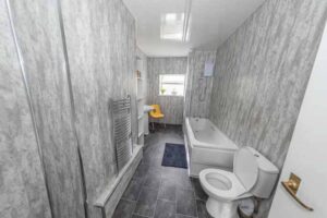Property for sale A modern bathroom in a traditional public house conversion, featuring gray marble-style walls and floor, a bathtub with shower, white toilet, towel radiator, small window, yellow chair, and blue rug—located in the Newcastle area. Presented by Palace Auctions
