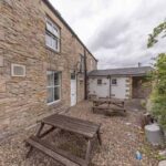 Property for sale The image shows the beer garden of a detached public house for sale in Burnopfield, Newcastle upon Tyne, with wooden picnic tables on gravel beside a stone building and white doors. Some greenery is visible to the right under a cloudy sky. Presented by Palace Auctions