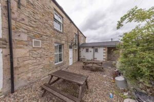 Property for sale The image shows the beer garden of a detached public house for sale in Burnopfield, Newcastle upon Tyne, with wooden picnic tables on gravel beside a stone building and white doors. Some greenery is visible to the right under a cloudy sky. Presented by Palace Auctions