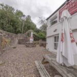 Property for sale A gravel outdoor seating area with picnic tables and large white umbrellas sits next to a detached public house for sale in Burnopfield, Newcastle upon Tyne, bordered by stone walls and trees under a cloudy sky. Presented by Palace Auctions