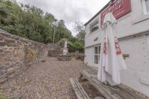 Property for sale A gravel outdoor seating area with picnic tables and large white umbrellas sits next to a detached public house for sale in Burnopfield, Newcastle upon Tyne, bordered by stone walls and trees under a cloudy sky. Presented by Palace Auctions
