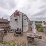 Property for sale Outdoor seating area with wooden benches and tables beside a stone building marked "Petton Arms." This detached public house for sale in Burnopfield, Newcastle upon Tyne offers a freehold property with beer garden, surrounded by stone walls and houses. Presented by Palace Auctions
